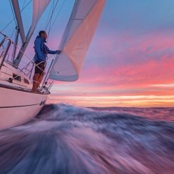 Man stading at the side of the deck of a yacht holding the jib while looking at the sunset